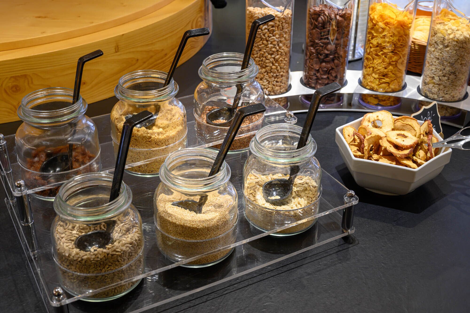 Variedad de alimentos en frascos y recipientes, incluyendo cereales y frutas secas, exhibidos sobre una barra.