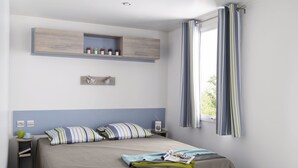 A bedroom with a bed, a nightstand, a shelf with decorative items, a window with curtains, and a green rug. - Marielyst Feriepark & Camping (Vaeggerlose)
