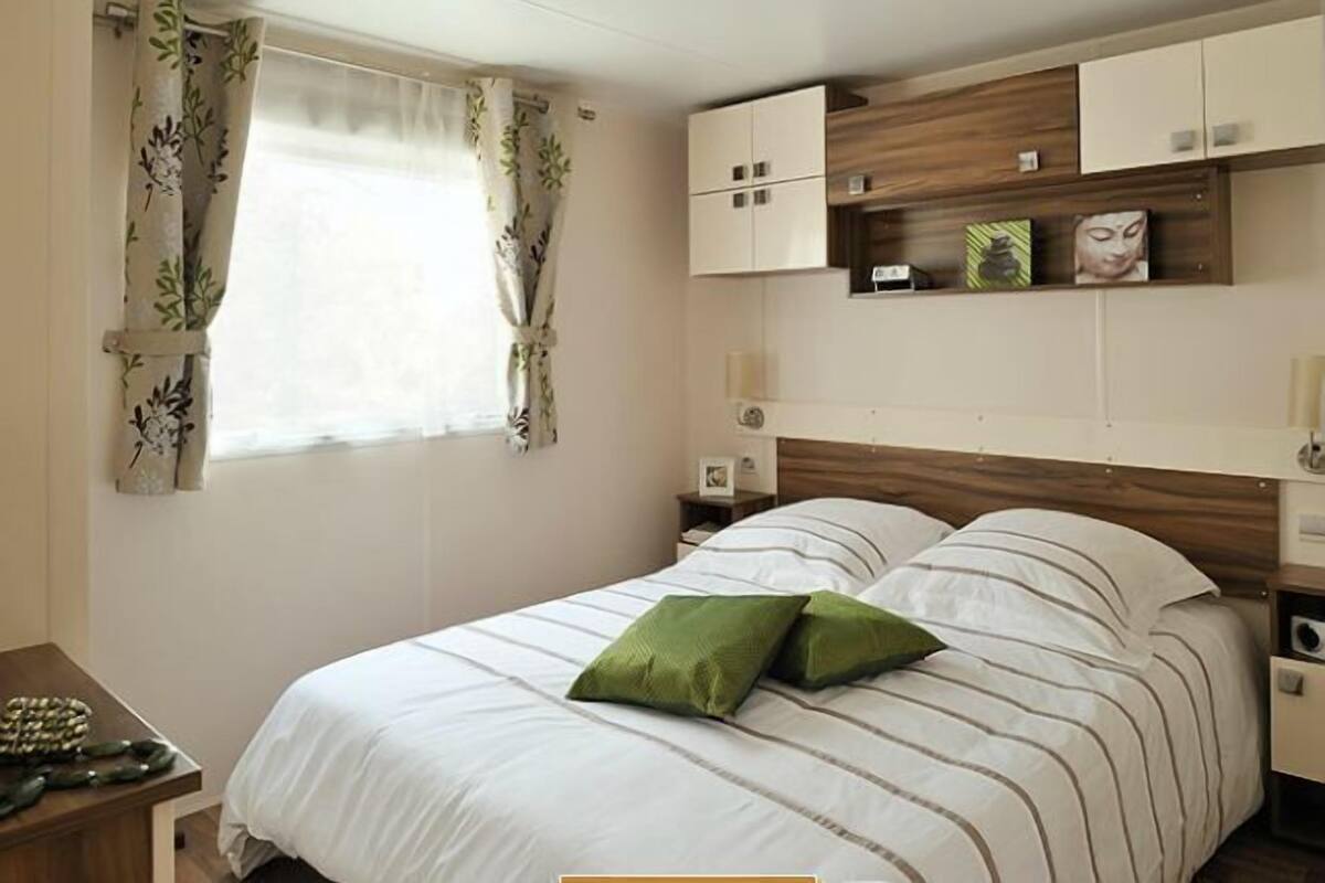 Ein ordentlich eingerichtetes Schlafzimmer mit einem Bett, grünen Kissen, einem Holz-Kopfteil und einem Fenster mit floralem Vorhang.
