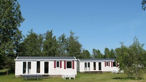 Two white mobile homes with red shutters, a bench, and a picnic table in a grassy field with trees in the background. - Marielyst Feriepark & Camping (Vaeggerlose)