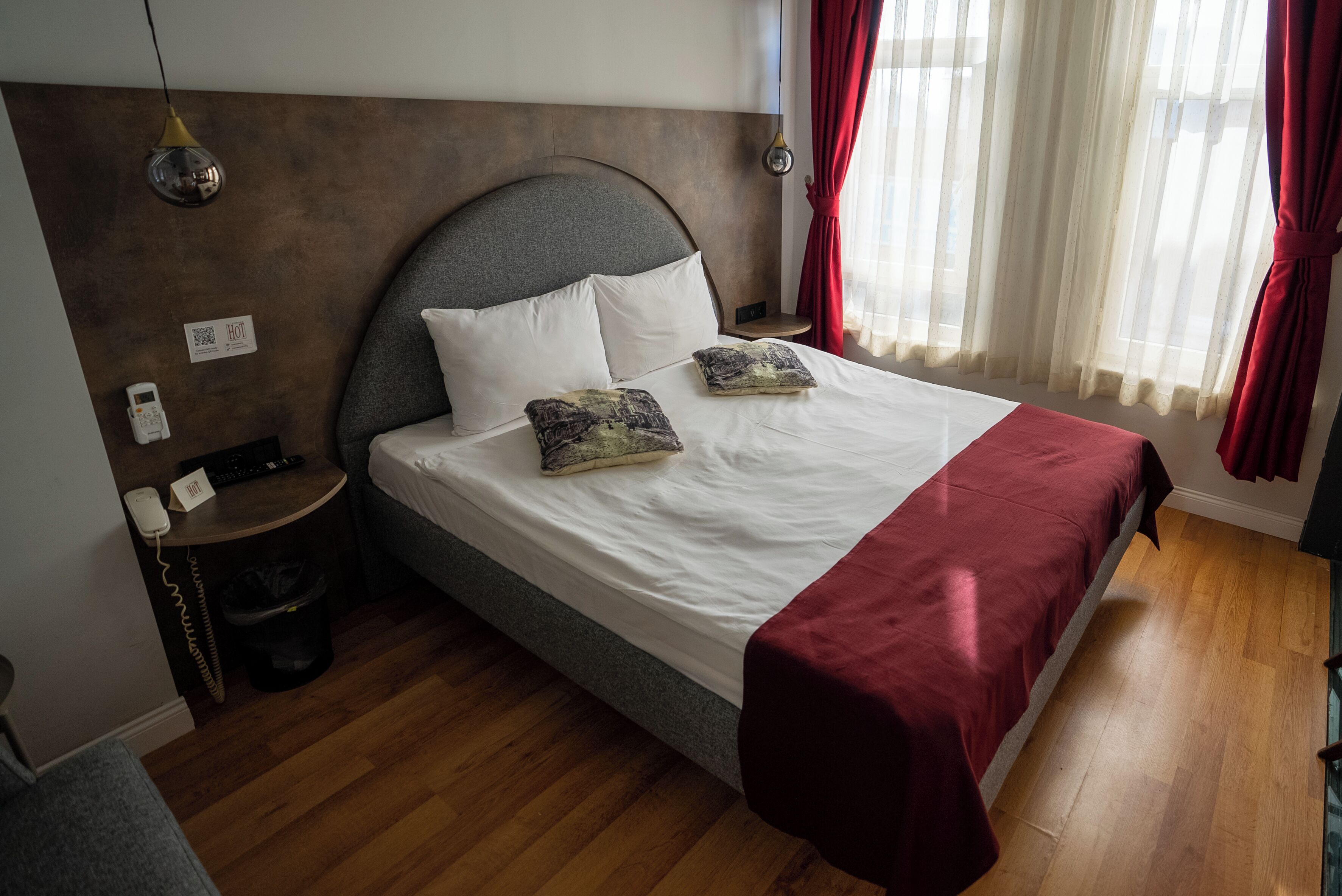 Una camera d'albergo con un letto grande, un comodino con telefono e cestino, una lampada a parete e una finestra con tende rosse.