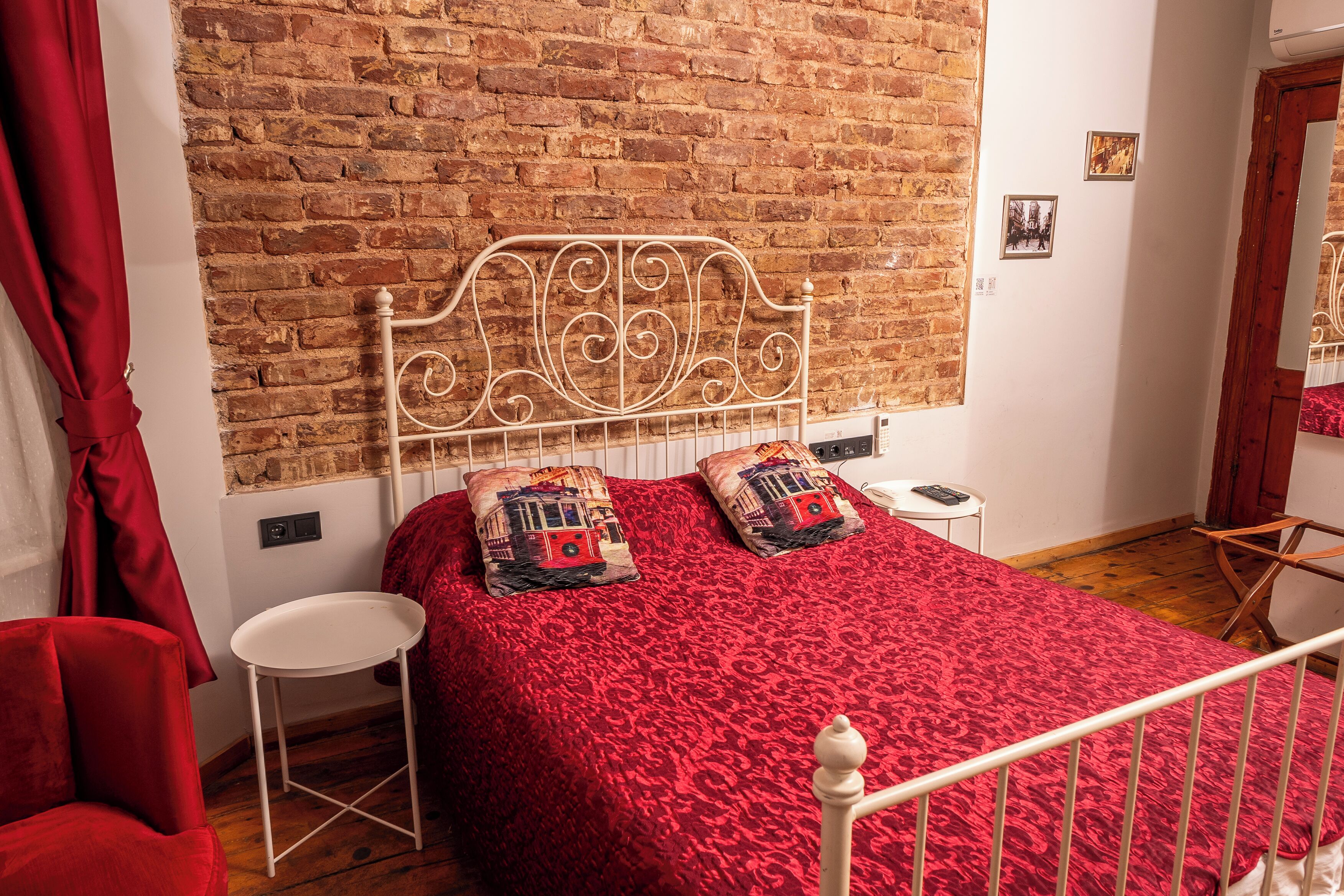 A bedroom with a brick wall, a white metal bed frame, a red bedspread, two matching pillows, a small white table, and a red chair.