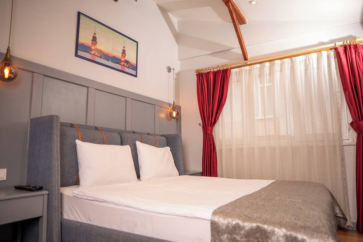 Habitación de hotel con cama, mesita de noche, un cuadro de una torre y cortinas rojas.