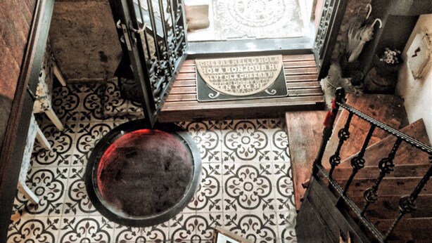 Una escalera con barandillas metálicas ornamentadas, baldosas de suelo estampadas y un candelabro con adornos de cristal.