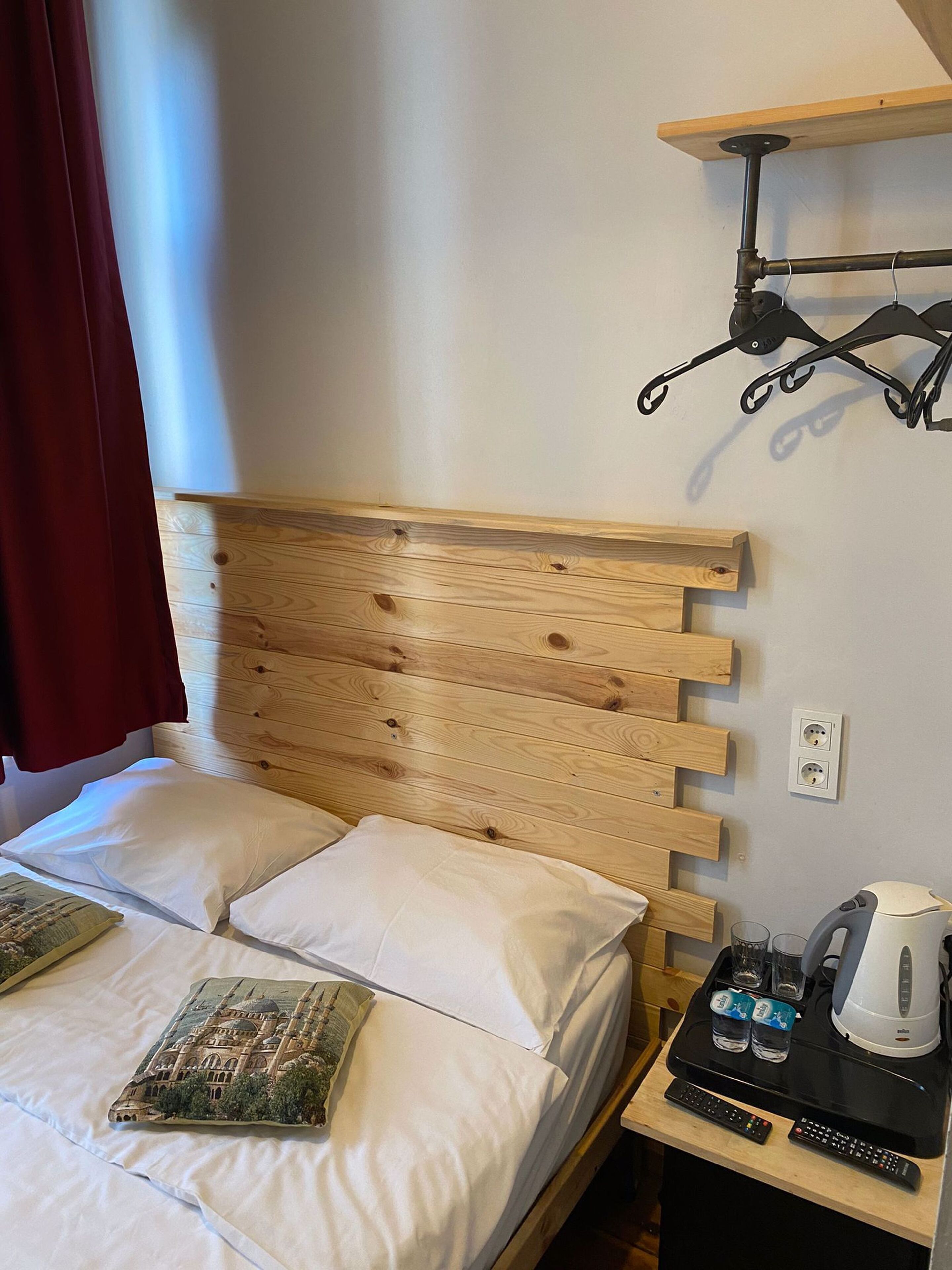 A wooden bed with white linens, a small bedside table with a kettle, glasses, and remote controls.