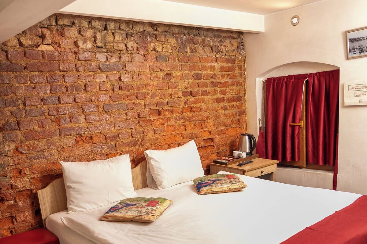 Un dormitorio con pared de ladrillo, cama de madera, banca roja, cortina roja y una pequeña mesa con un hervidor.