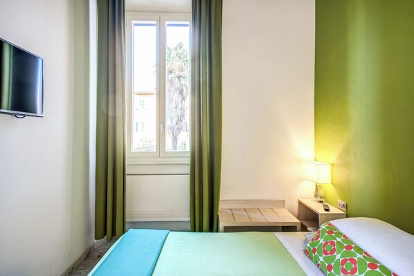 Une chambre d’hôtel avec un lit, un téléviseur fixé au mur, une fenêtre avec des rideaux, une table de chevet avec une lampe, et une vue sur l’extérieur.