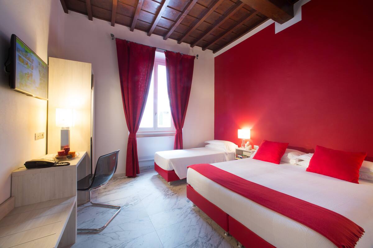 A hotel room with two beds, a desk with a chair, a lamp, and a window with red curtains.