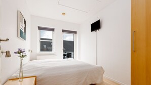 A bedroom with a bed, a bedside table with a remote control, a vase with flowers, and a window with a view of a building. - Hotel Rudkøbing Skudehavn (Rudkobing)