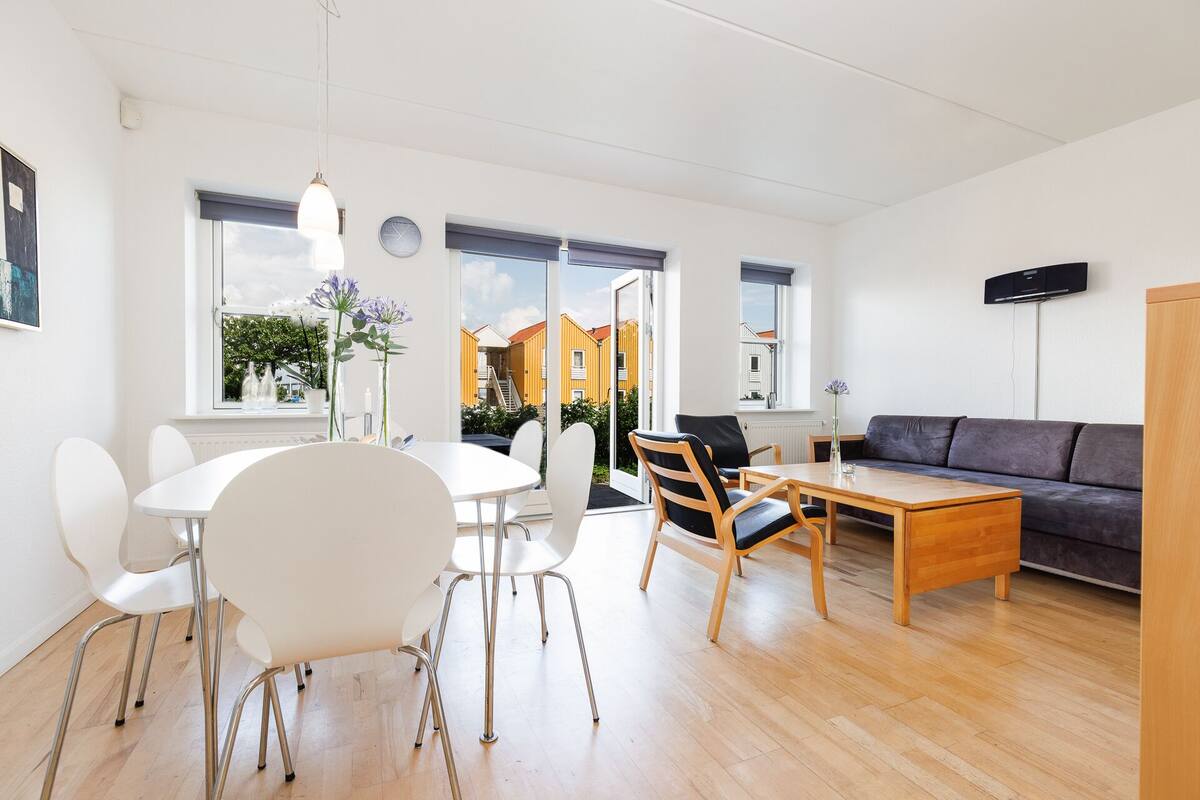 Una sala de estar moderna con comedor, un sofá y un televisor. Hay ventanas amplias con vistas a edificios.