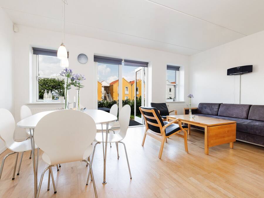 A modern living room with a dining area, a sofa, and a TV. There are large windows with a view of buildings outside.