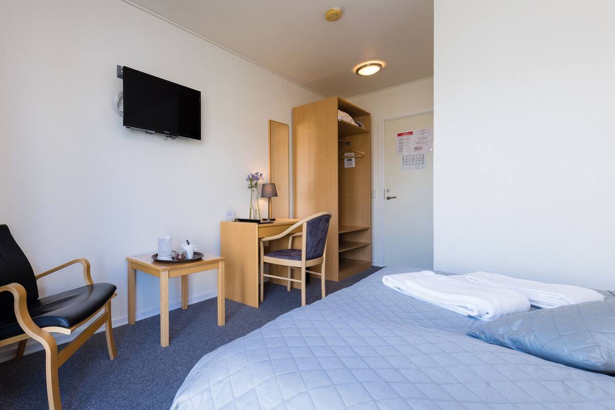 Habitación de hotel con una cama, un escritorio con silla, un televisor montado en la pared y un armario.
