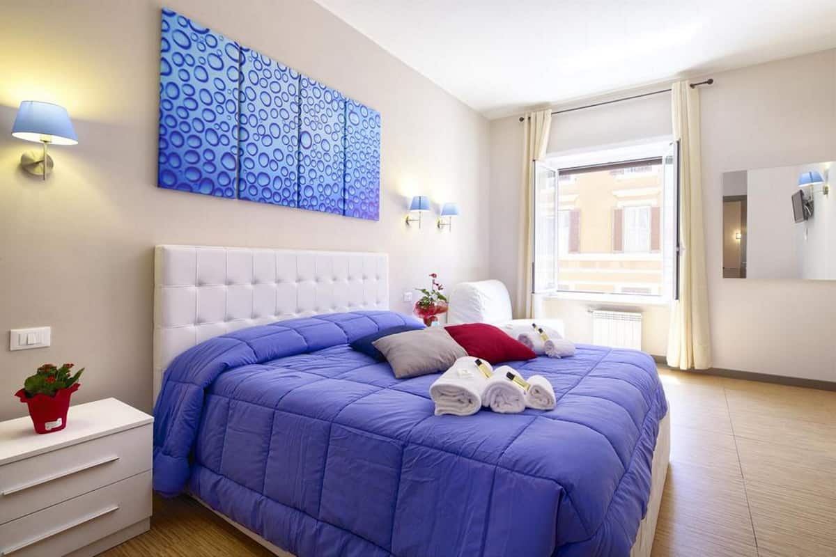 A modern hotel room with a large bed, a white headboard, a blue abstract painting, and a view of the city through the window.