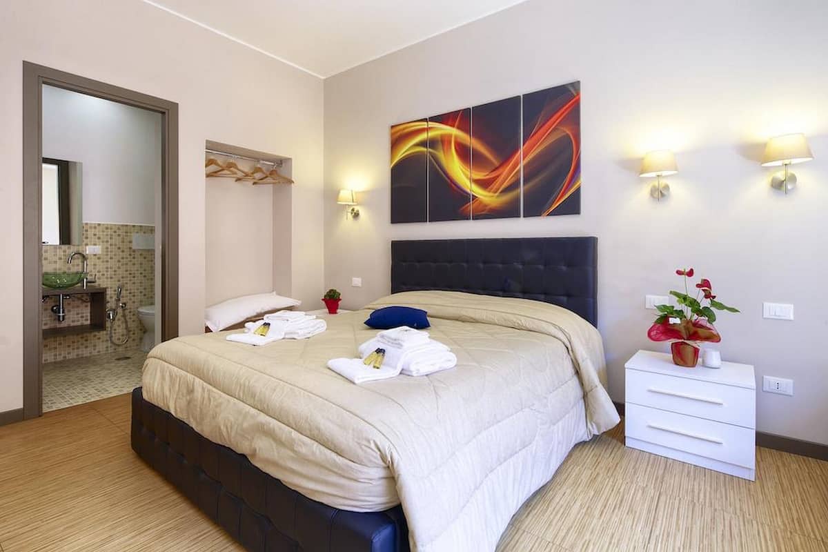 A hotel room with a large bed, a bedside table with a lamp, a wall-mounted artwork, and a bathroom visible through an open door.