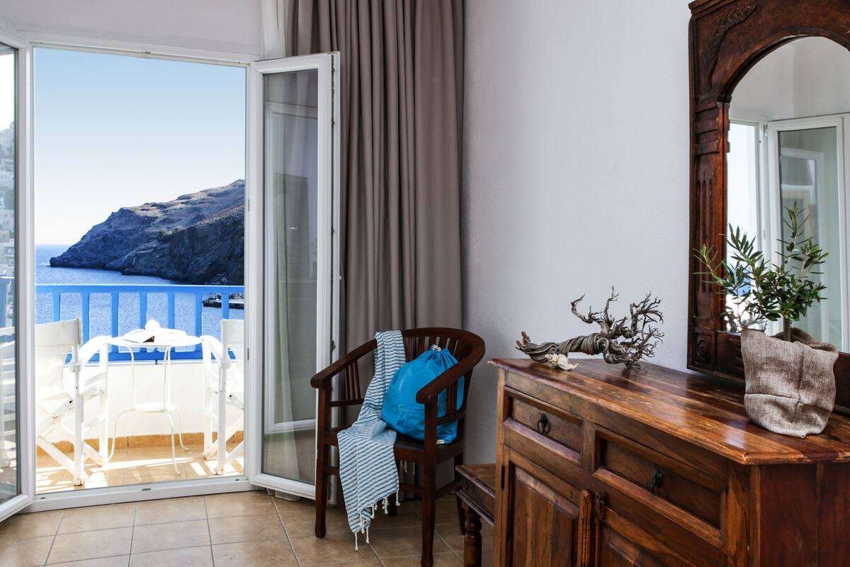 A room with a wooden dresser, a chair with a blue towel, and a view of the sea through a sliding glass door.
