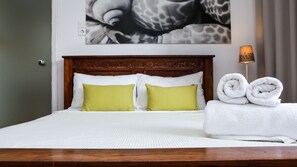 Ein ordentlich bezogenes Bett mit weißer Bettwäsche und zwei grünen Kissen, ein Holz-Kopfteil, eine Wandlampe und ein aufgerolltes weißes Handtuch auf der Seite.