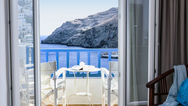 A balcony with white chairs and a table, overlooking a rocky coastline and the sea. - Akti Rooms (Astypalaia)