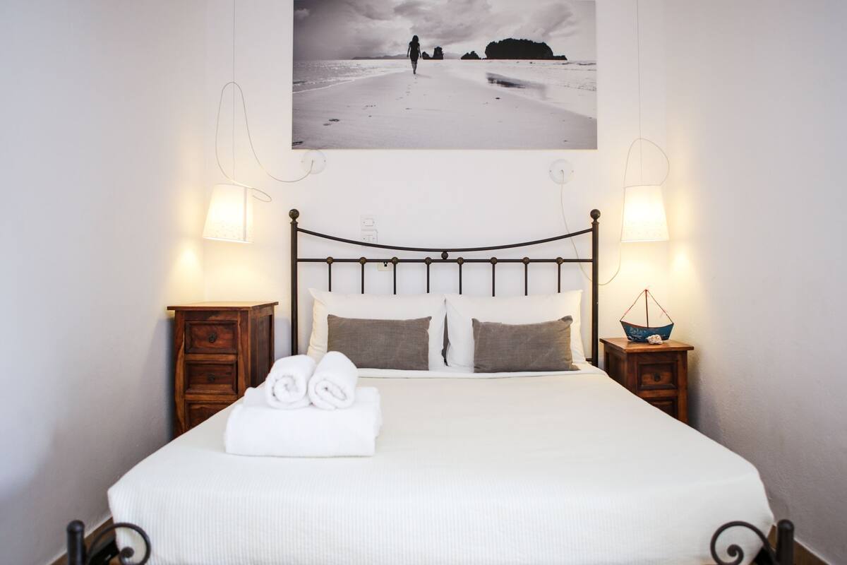 A neatly made bed with a rolled towel, two bedside tables with lamps, and a framed picture of a beach scene above the bed.