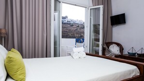A hotel room with a bed, a chair, a TV, and a view of a coastal town. - Akti Rooms (Astypalaia)