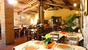 A restaurant with wooden beams and brick walls, tables set with orange tablecloths and flower centerpieces. - Fattoria Il Santo (Monticiano)