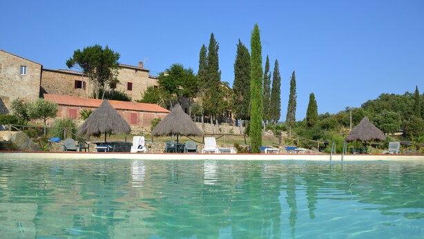 A clear pool with lounge chairs and umbrellas, a stone building with red shutters, and tall cypress trees. - Fattoria Il Santo (Monticiano)