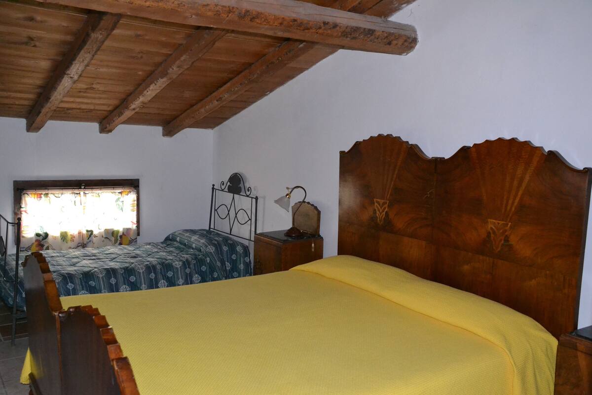 Een slaapkamer met een houten plafond, een bed met een gele sprei, een houten hoofdbord en een raam met gordijnen.
