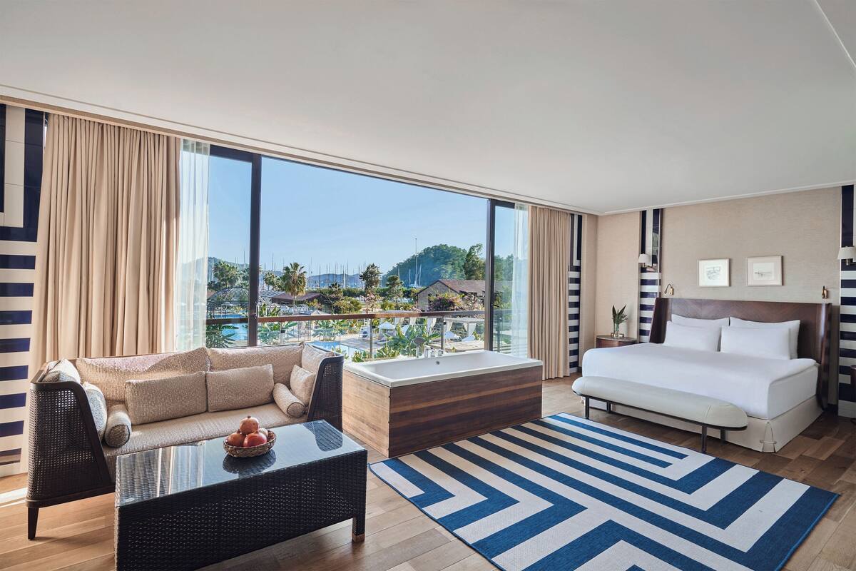 Une chambre d’hôtel moderne dotée d’un grand lit, d’un canapé, d’une table basse et offrant une vue sur un balcon aménagé avec du mobilier d’extérieur.