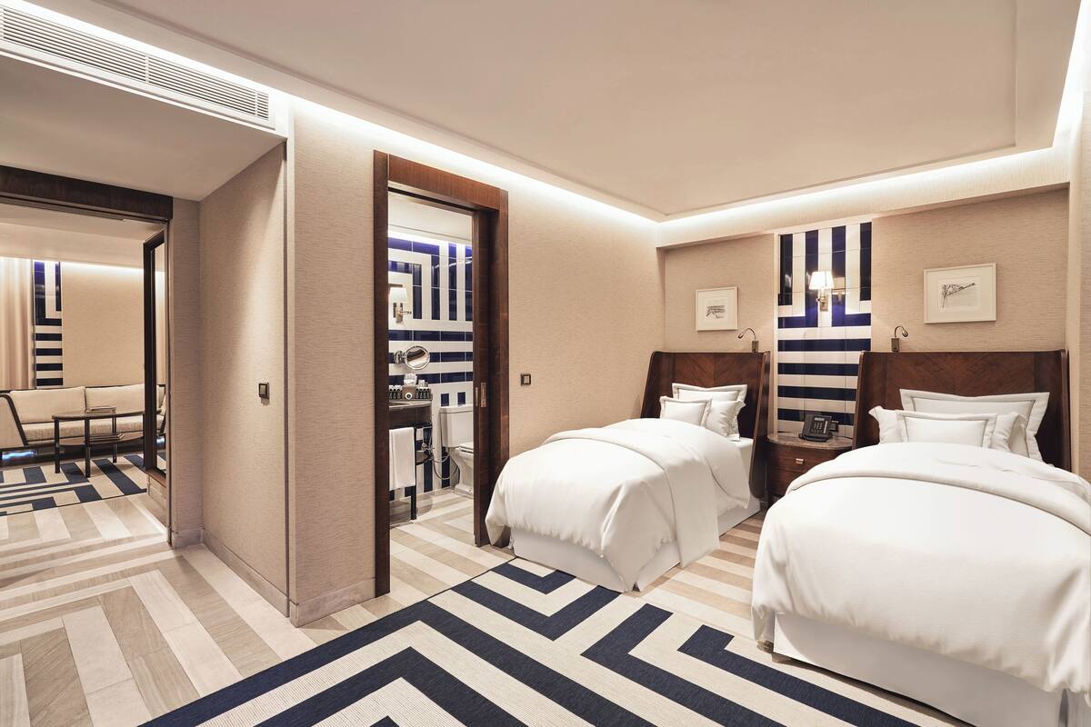 Une chambre d’hôtel avec deux lits, un canapé, un miroir et une salle de bain visible à travers une porte ouverte.