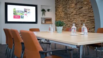 A conference room with a long table, orange chairs, a projector screen, a wooden wall, and a potted plant.