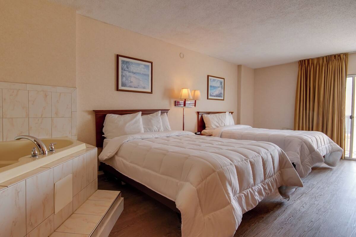 Habitación de hotel con una bañera grande, dos camas con ropa blanca, un cabecero de madera y un cuadro en la pared.