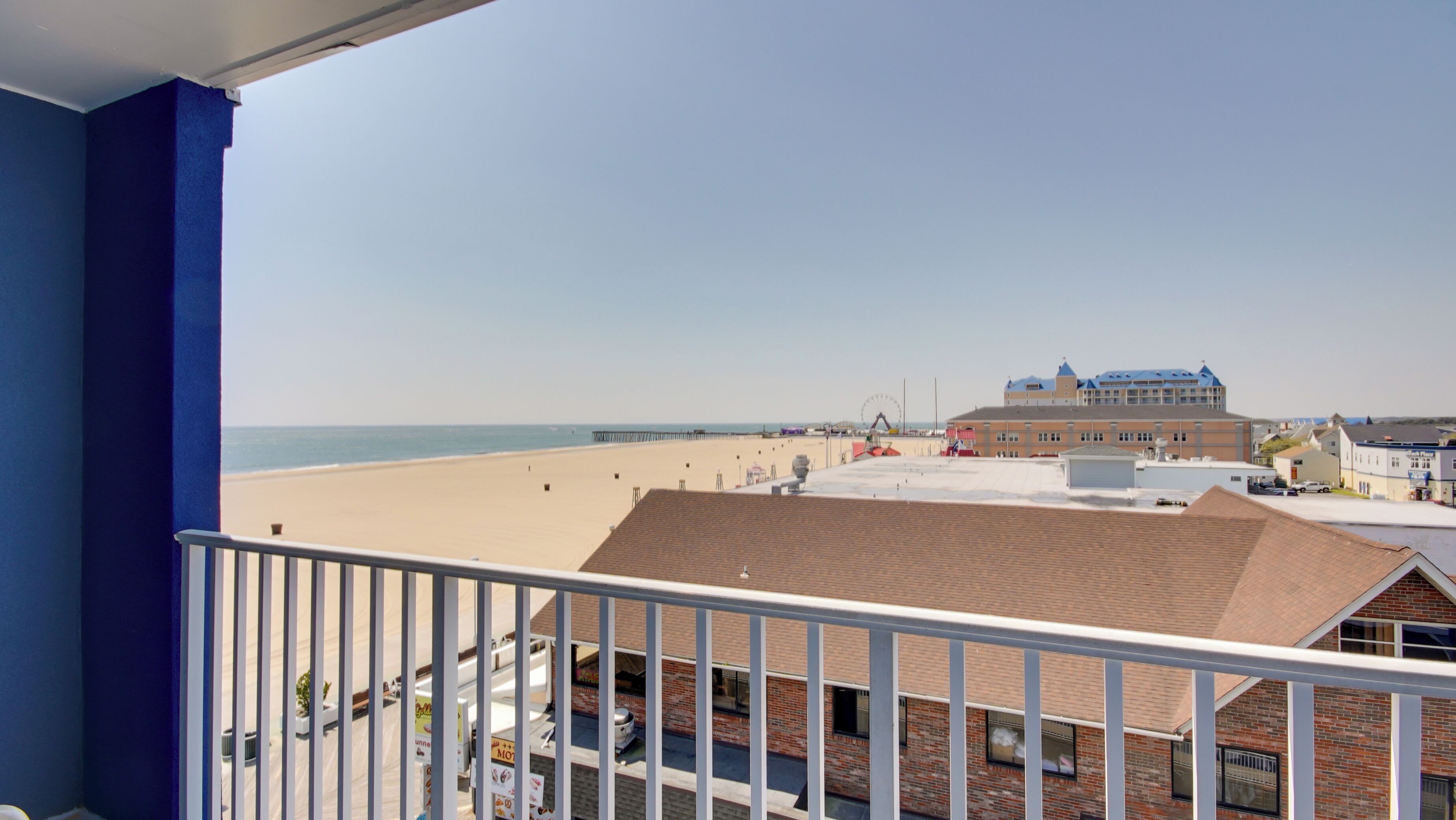 A beach view from a balcony with a clear sky, a sandy beach, and a building with a blue roof in the background.