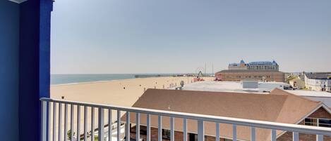 A beach view from a balcony with a clear sky, a sandy beach, and a building with a blue roof in the background.