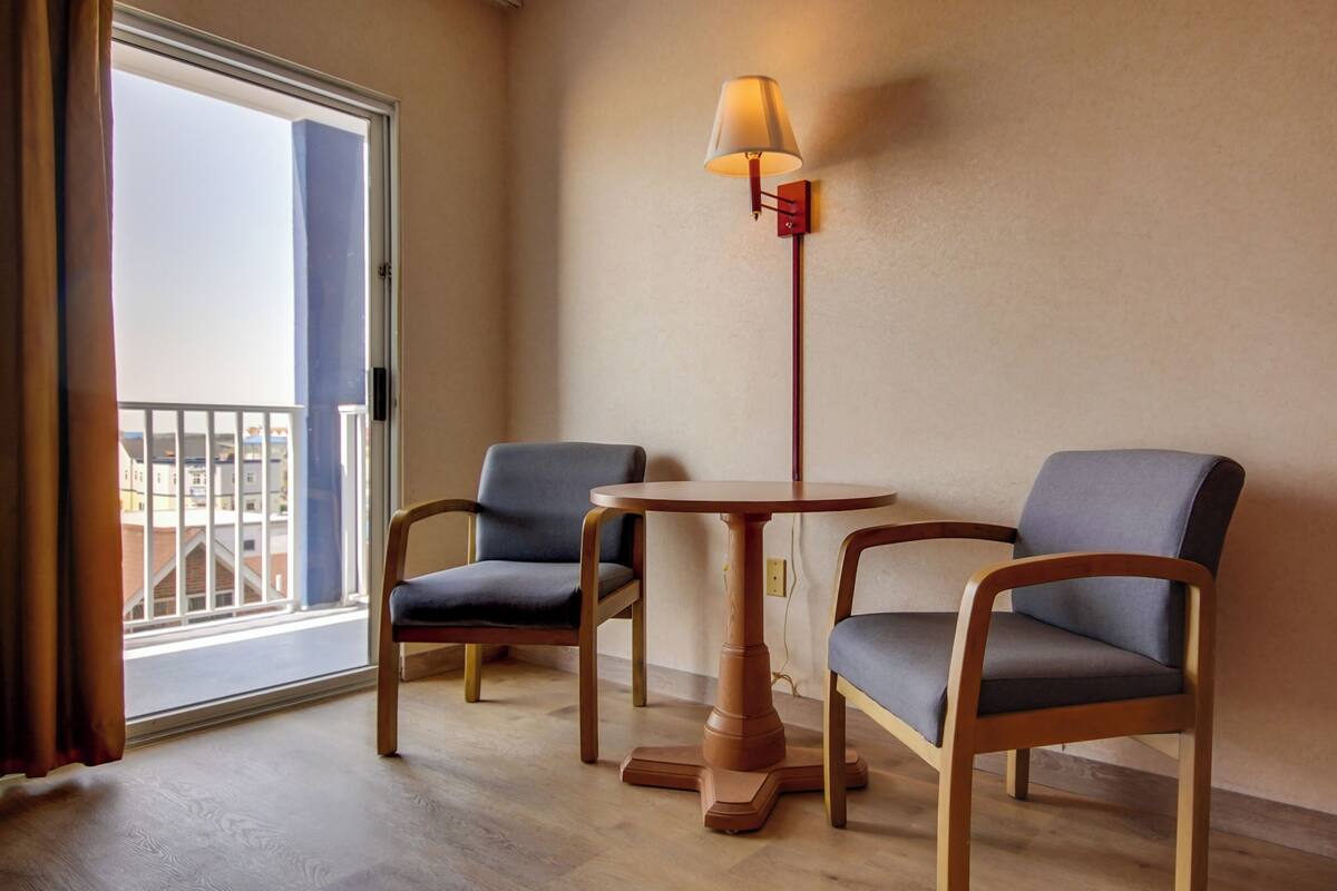Una habitación con dos sillas, una mesita pequeña, una lámpara y vistas a edificios a través de una ventana.