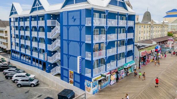 Un hotel de varios pisos con balcones, un paseo marítimo con tiendas y un estacionamiento.