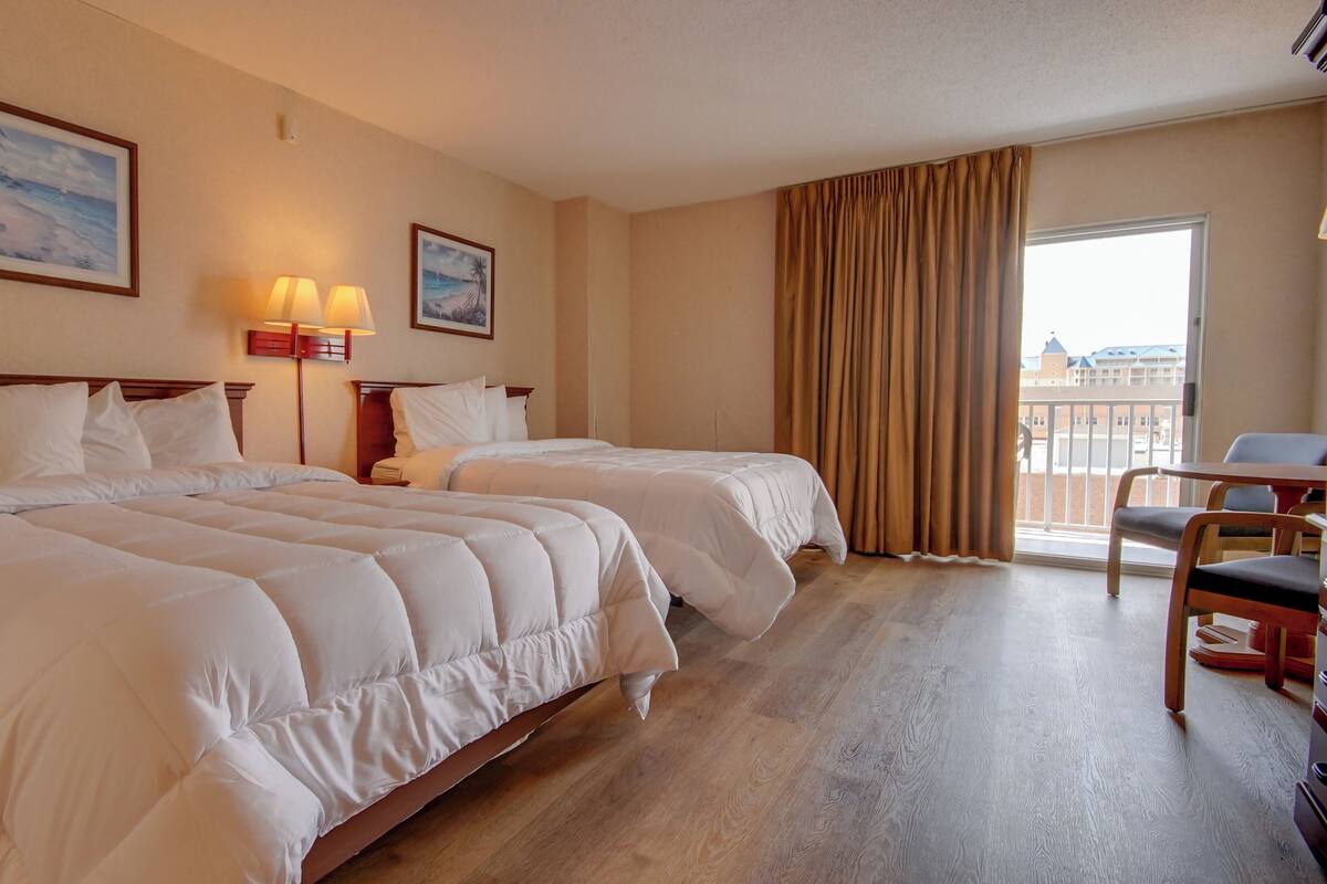 Habitación de hotel con dos camas, un escritorio, una silla, un balcón con cortinas y dos lámparas de pared.