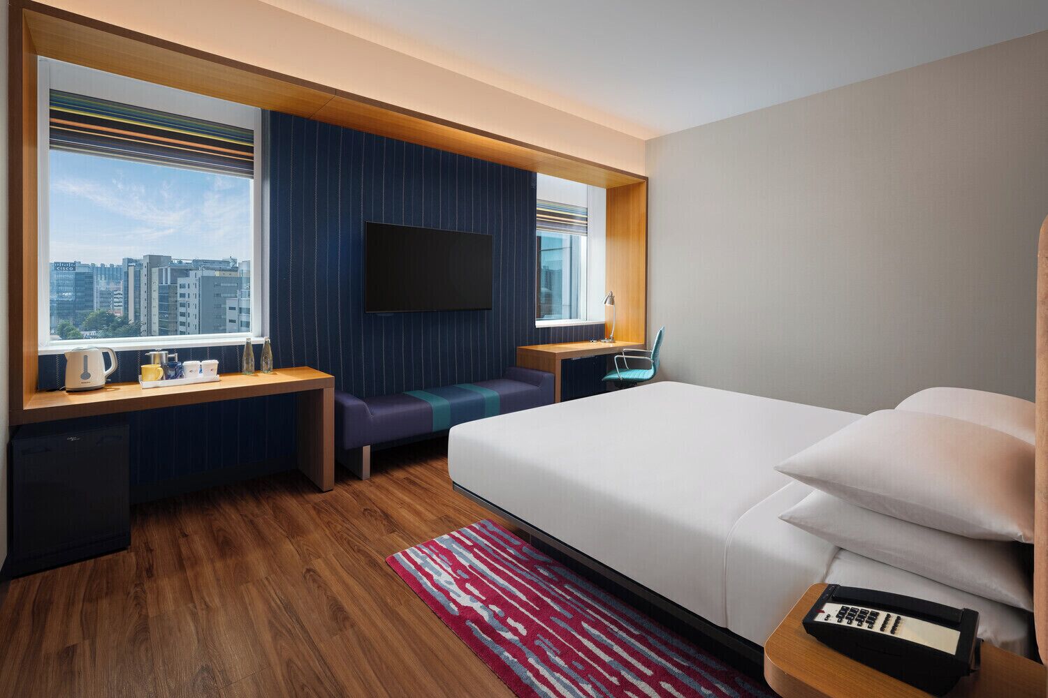 Habitación de hotel con cama, escritorio, televisor, ventana con vista a la ciudad y alfombra roja con diseño.