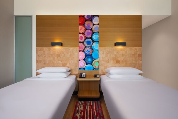 A hotel room with two single beds, a wooden headboard with a colorful circular design, a small nightstand with a phone, and a red and beige striped rug.