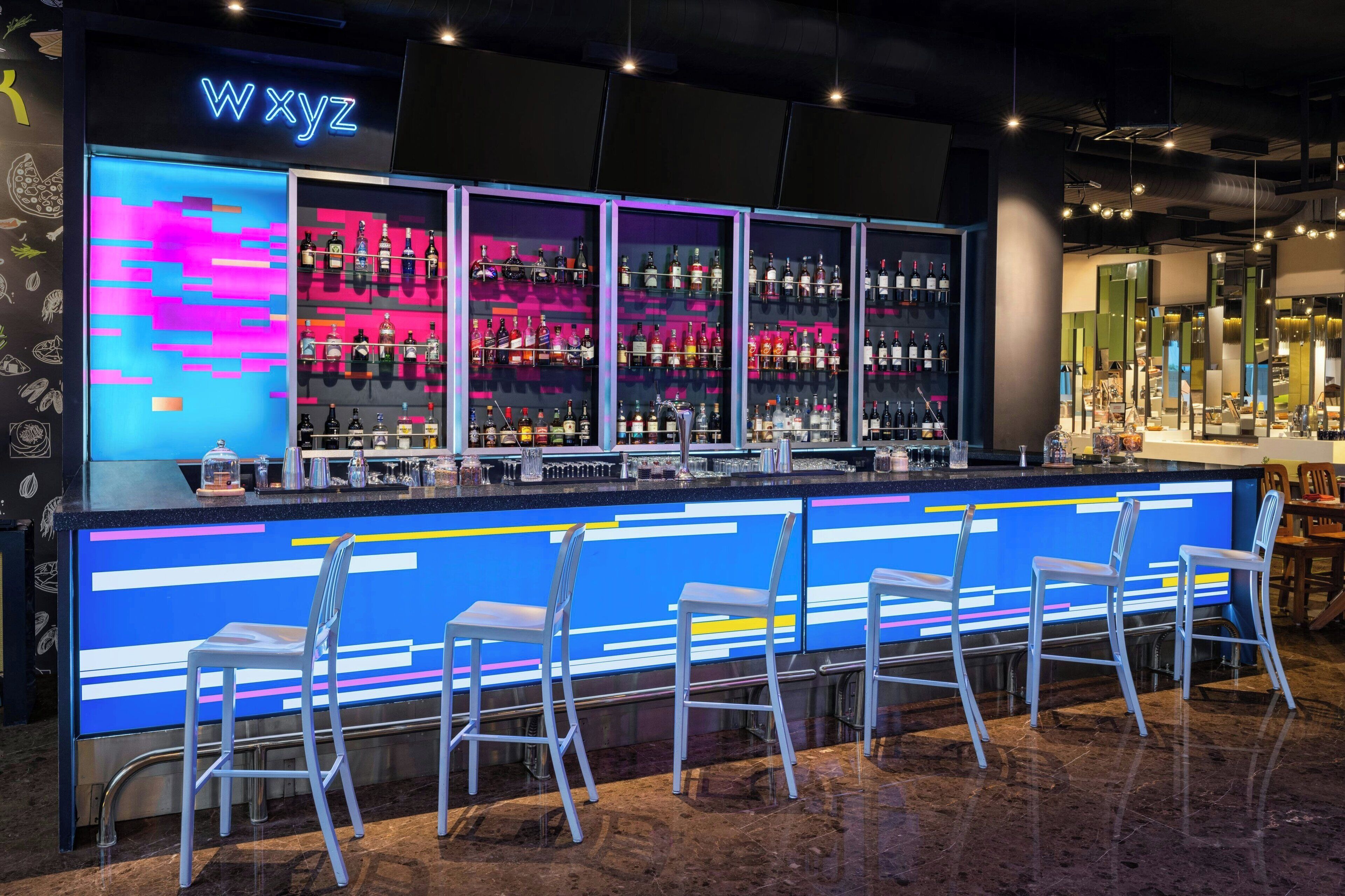 A bar area with a neon sign reading "WXYZ," multiple shelves of liquor bottles, and four bar stools.
