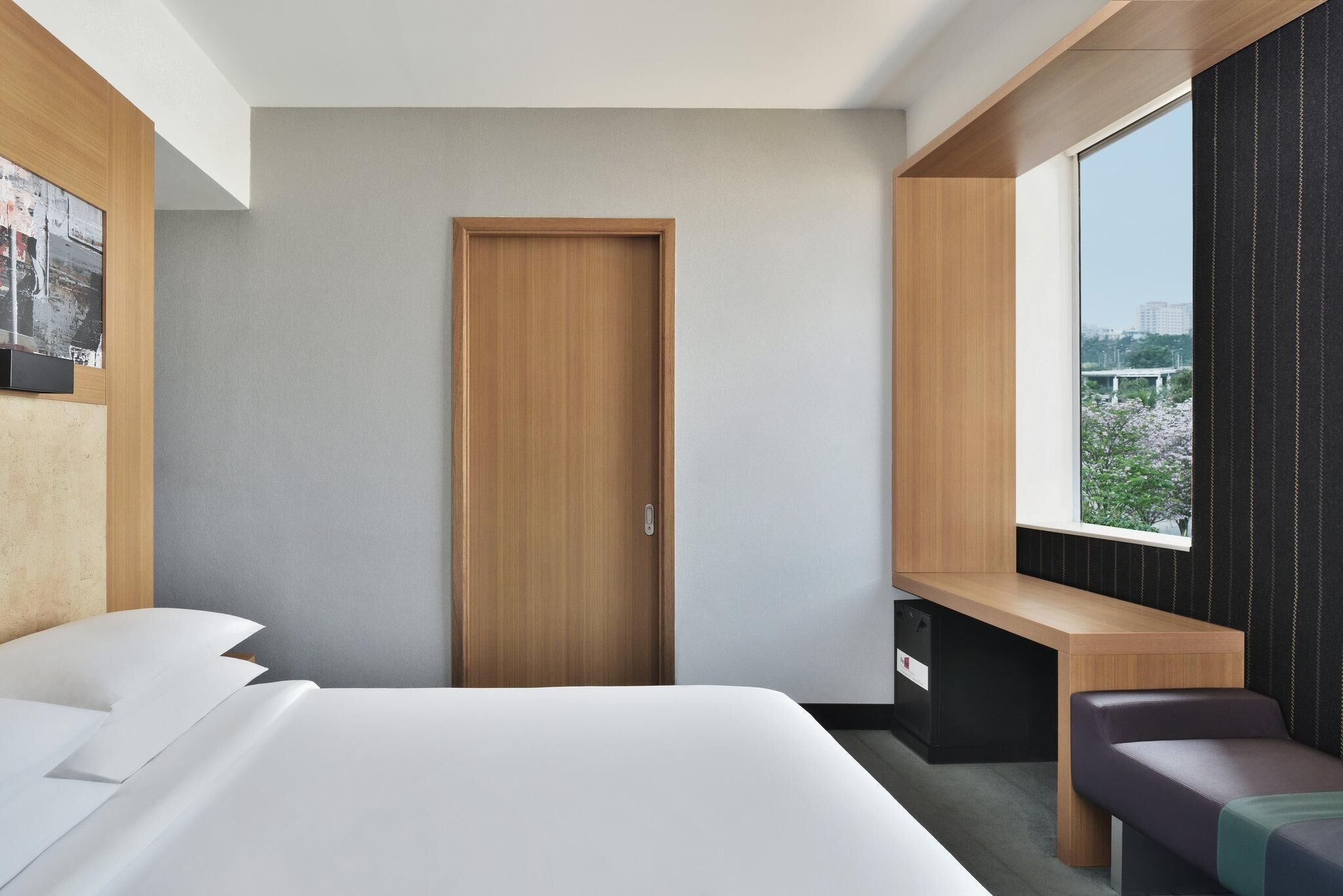A modern hotel room with a bed, a desk, a bench, a door, and a large window offering a city view.