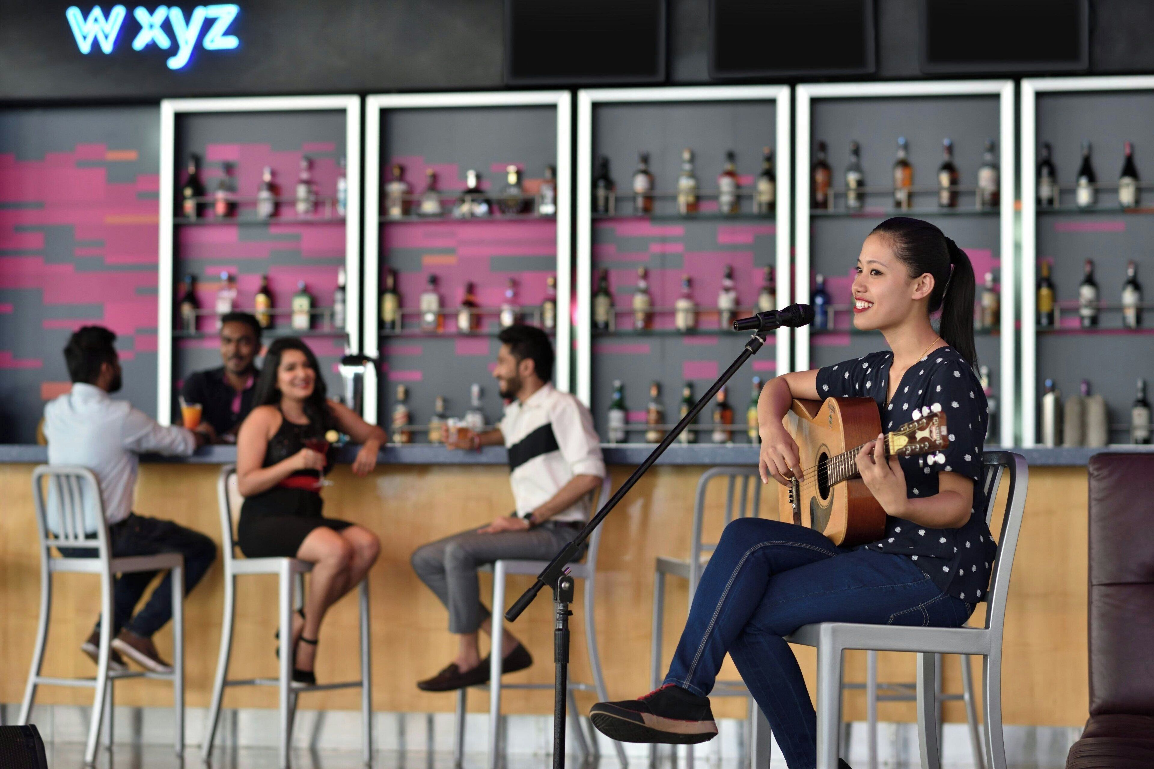 A bar area with a microphone stand and a person playing guitar. There are chairs and people sitting at the bar.