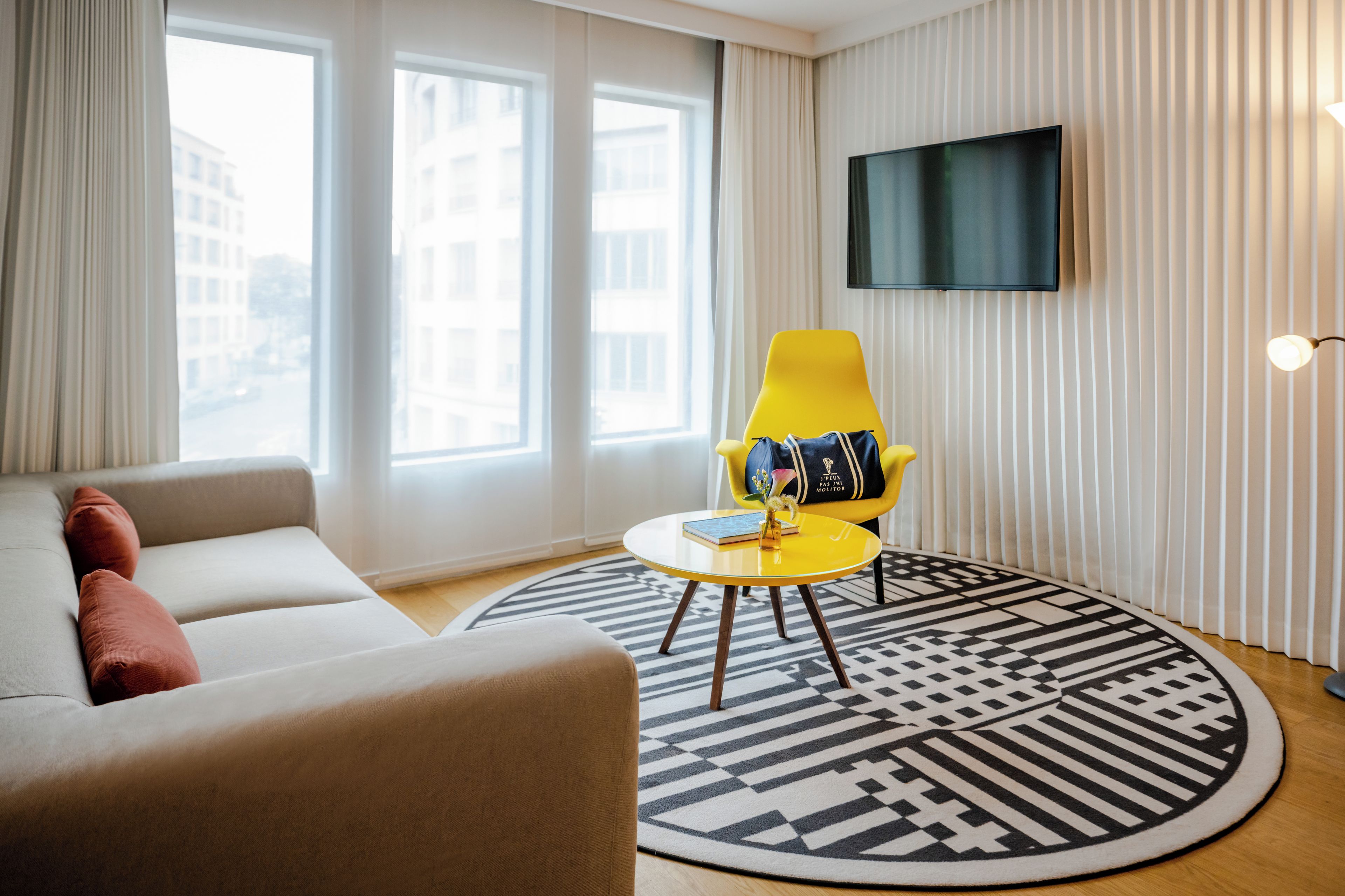 A modern living room with a sofa, a yellow armchair, a small table with a book and a vase, and a TV mounted on the wall.