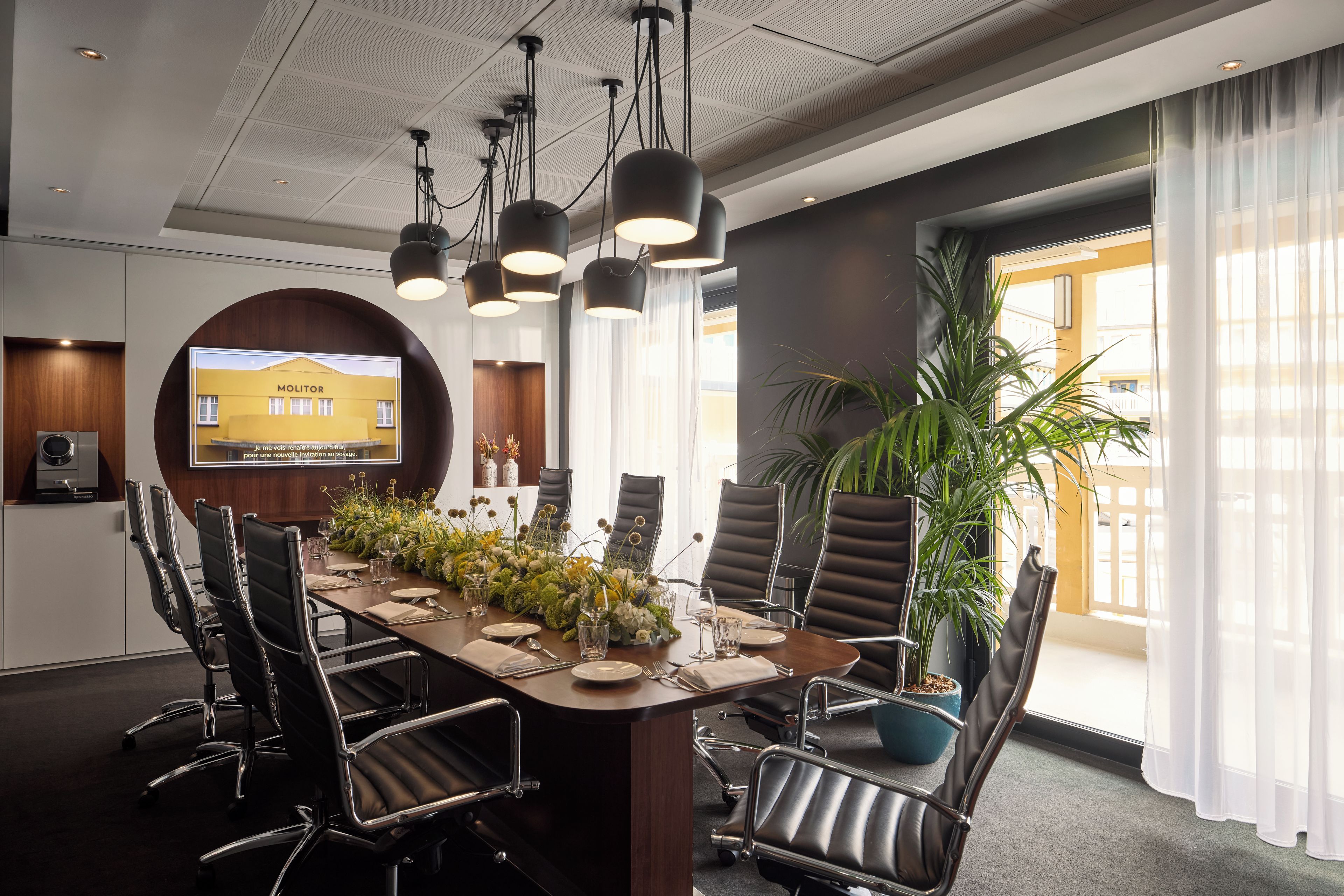 Ein Konferenzraum mit einem langen Tisch, Stühlen, einer Projektionsleinwand und einem Blumentopf.