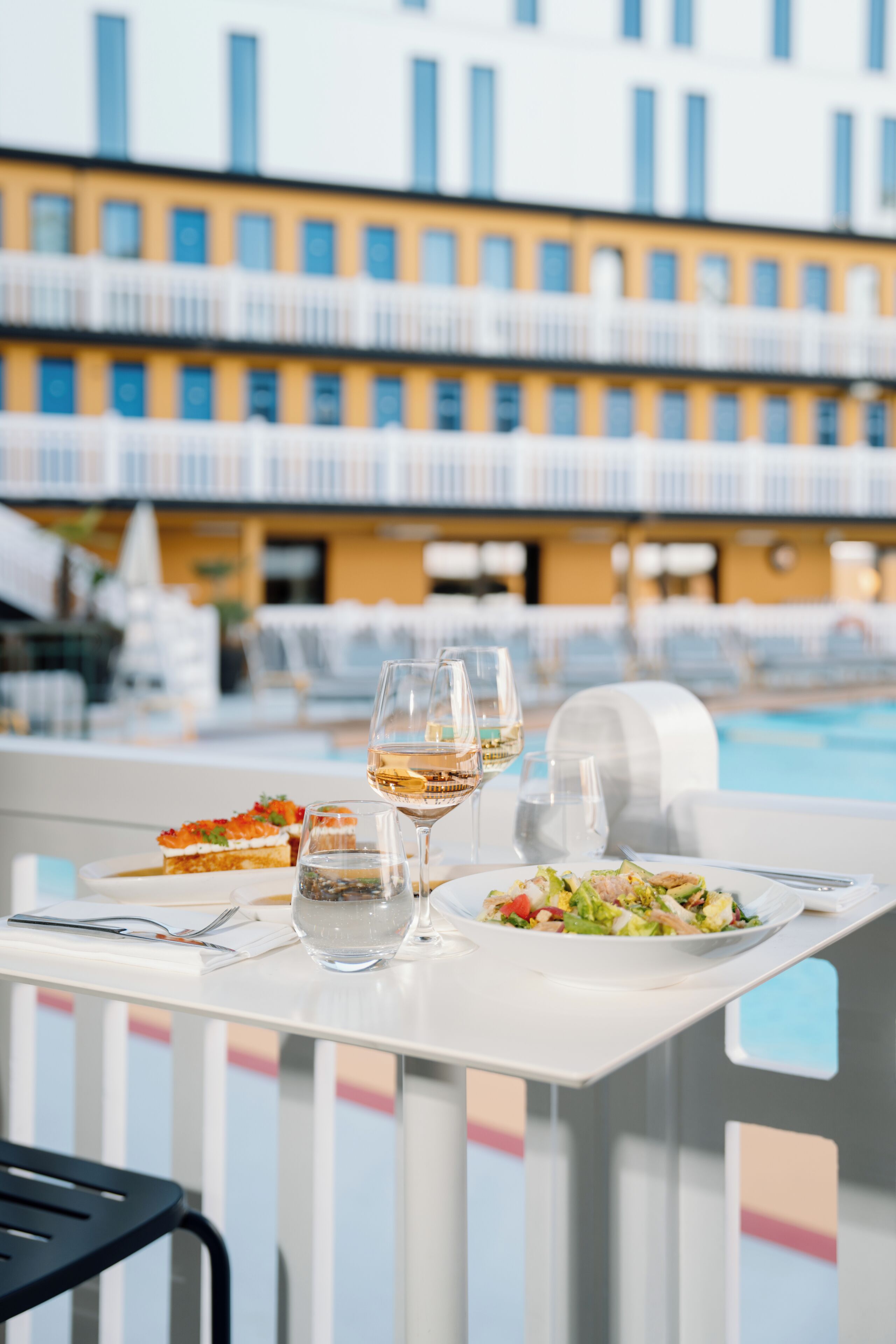 A dining table with a meal, glasses, and a poolside view.