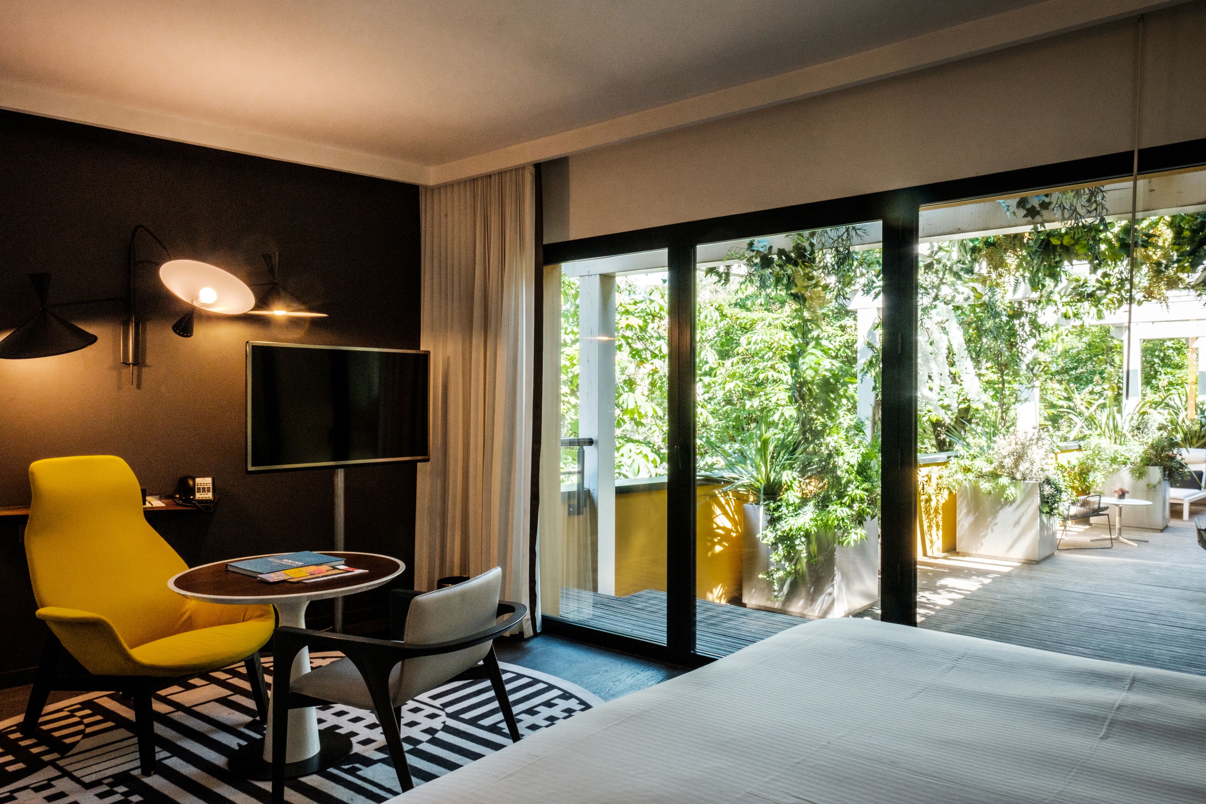 Ein Hotelzimmer mit einem Bett, einem gelben Sessel, einem kleinen Tisch, einem Fernseher und einem Balkon mit Pflanzen und Blick ins Grüne.