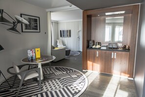 A modern hotel room with a round dining table, a sofa, a kitchen area with cabinets, a flat-screen TV, and a patterned rug.