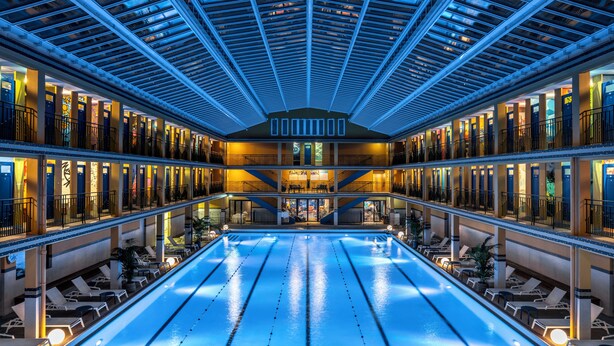 Una amplia piscina interior con techo de cristal, varios niveles de balcones y zonas de descanso con tumbonas.