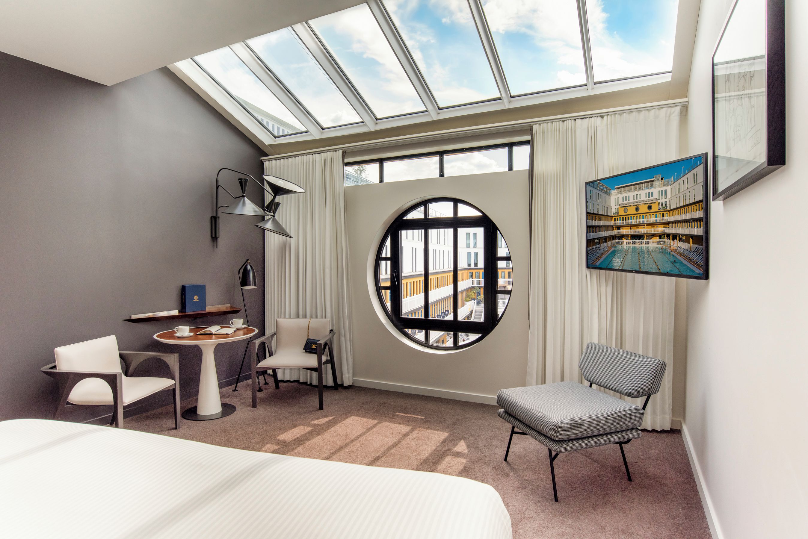 Een hotelkamer met een dakraam, een rond raam, een bed, een bureau, twee stoelen en een schilderij van een gebouw.