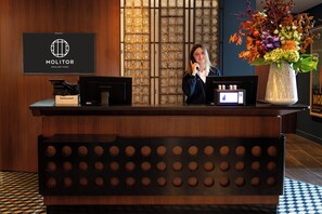 A reception desk with a logo, a computer, a phone, and a vase with flowers. - Molitor Hotel & Spa Paris - MGallery Collection (Paris)