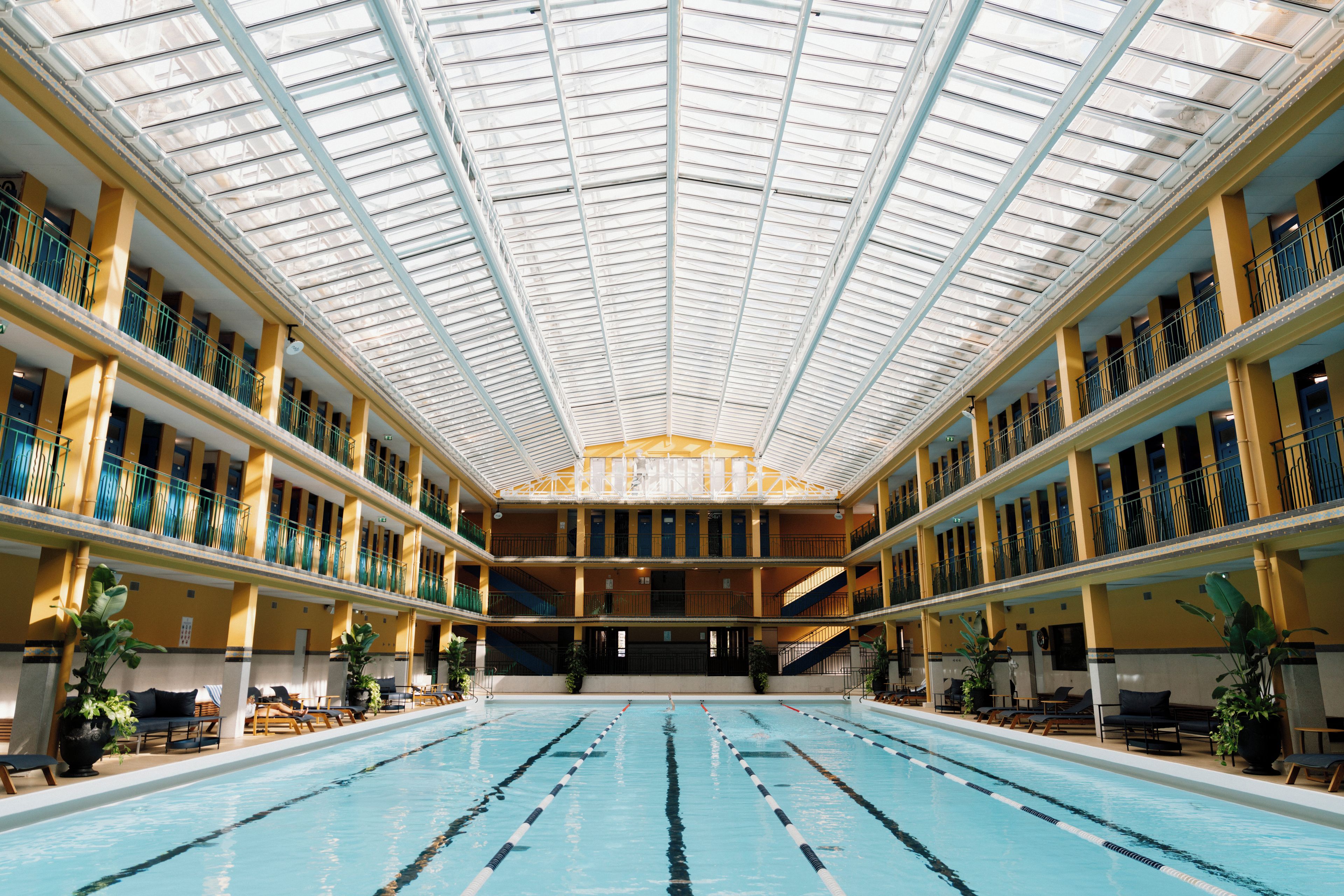 Une grande piscine intérieure dotée de plusieurs couloirs, entourée de balcons avec des garde-corps et surmontée d’un haut plafond agrémenté de lucarnes.