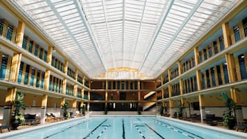 Une grande piscine intérieure dotée de plusieurs couloirs, entourée de balcons avec des garde-corps et surmontée d’un haut plafond agrémenté de lucarnes.